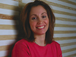 A smiling woman with brown hair wearing a pink top, standing in front of a tan and white striped wall.