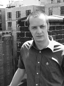 A man wearing a striped short-sleeved button-down shirt stands in front of brick buildings.