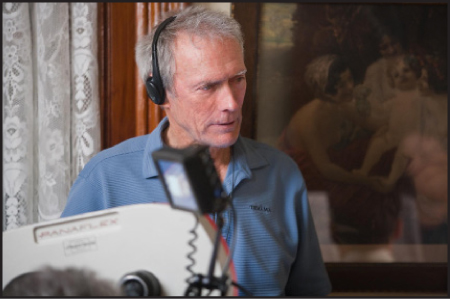 A man with grey hair and headphones looks intently at a monitor in a room with a painting.
