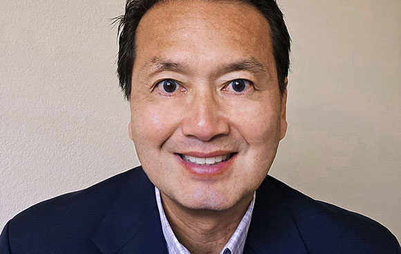 A smiling man with dark hair wearing a navy blazer and a light patterned shirt.