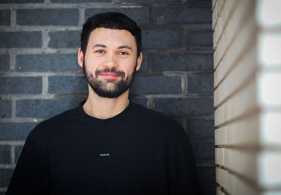 A man with a dark beard and short hair wearing a black t-shirt, smiling in front of a grey brick wall.