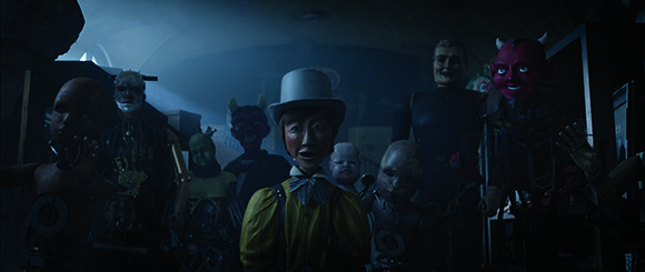 A group of eerie, vintage-style dolls and masks arranged in a dimly lit, shadowy room.