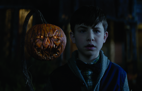 A young boy with a worried expression stands beside a large, scary carved pumpkin with glowing eyes.