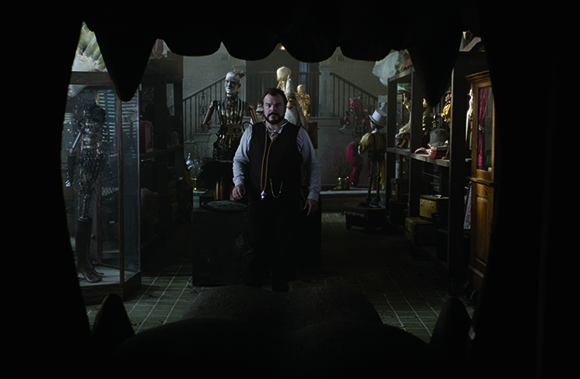 A man in a vest stands in a dimly lit room filled with mannequins and antique objects.