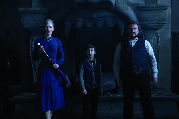 A woman in a blue dress, a young boy, and a man in a vest stand before a large stone monster-shaped fireplace.