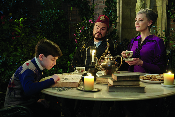 A boy reads a book while a man and woman sit at a candlelit table with tea and cookies.