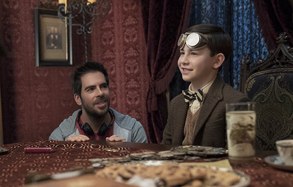 A man looks up at a smiling boy wearing aviator goggles and a bowtie at a dining table.