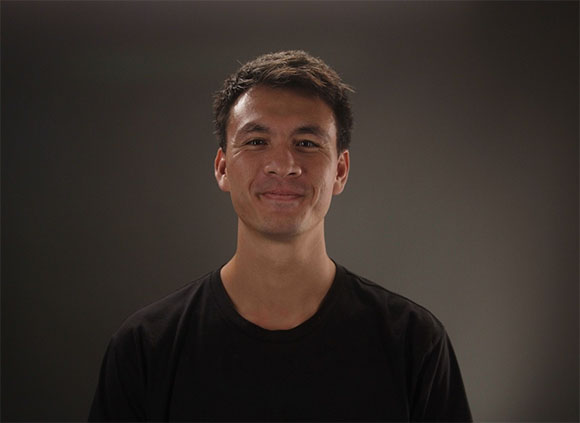 A man with dark hair and a slight smile wearing a black t-shirt against a dark gray background.