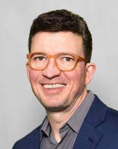 A man with short dark hair and orange-rimmed glasses smiling against a light gray background.