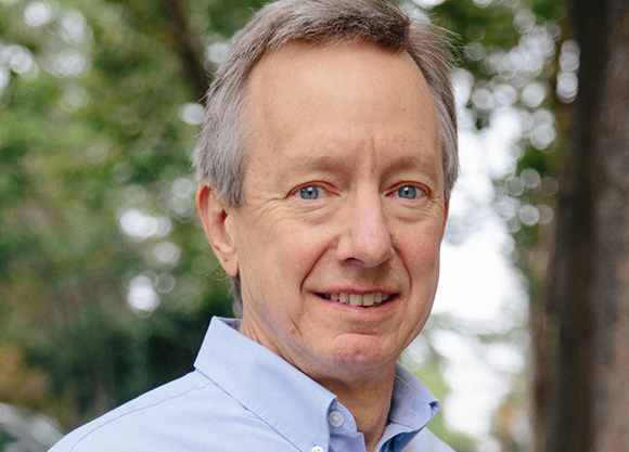 A middle-aged man with short graying hair and blue eyes smiling in a light blue button-down shirt.