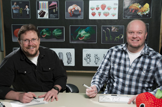 Two men sit at a desk with pens, positioned in front of a black board covered in various character sketches.
