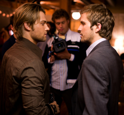 Two men in suits stand facing each other in a dimly lit room with a cameraman in the background.