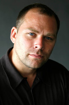 Close-up portrait of a man with short dark hair and blue eyes wearing a black polo shirt.