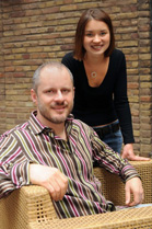 A man in a striped shirt sits in a wicker chair while a woman in a black top stands behind him.
