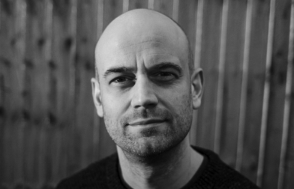 Black and white close-up portrait of a bald man with light facial stubble looking at the camera.