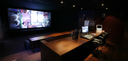 A dark control room featuring a large projection screen and a long wooden desk with computer monitors and equipment.