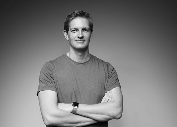 Black and white studio portrait of a man in a t-shirt standing with his arms crossed.