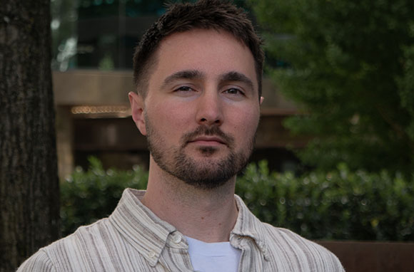 A man with short dark hair and a beard wearing a striped button-down shirt over a white t-shirt.