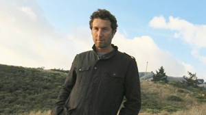 A man in a dark jacket stands outdoors against a backdrop of hills and a cloudy sky.