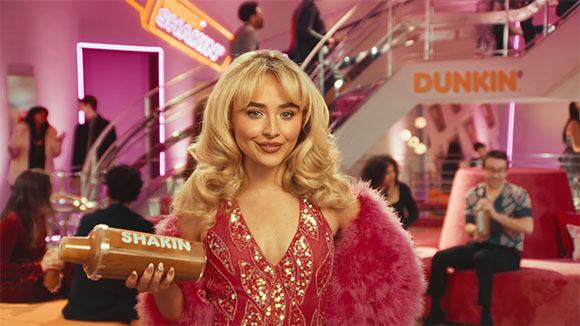 A blonde woman in a red sequined dress and pink fur shrug holds a shaker in a pink lounge.