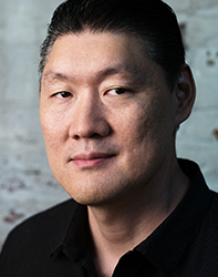 A man with dark hair and a black collared shirt looking toward the camera against a light brick wall.