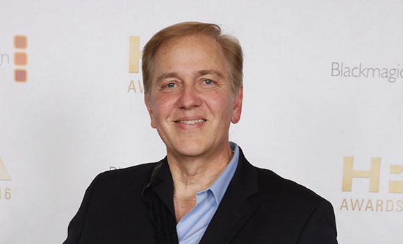 A man with light hair and a blue shirt smiling in front of a white backdrop with logos.