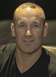 A close-up portrait of a man with short hair and a slight smile against a dark background.