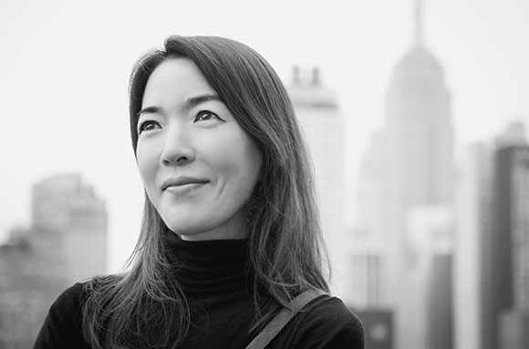 A woman with long dark hair looks upward with a slight smile against a blurred cityscape.