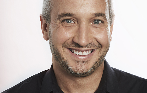 A smiling man with short graying hair and a beard wearing a black collared shirt against a white background.