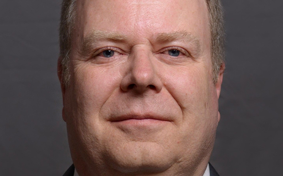 A close-up headshot of a middle-aged man with light skin and blue eyes looking forward.