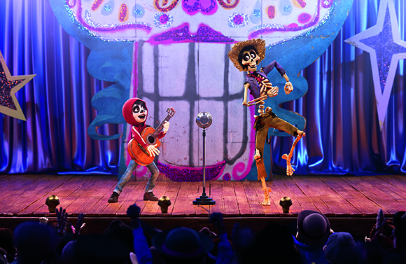 A skeleton playing a guitar and another dancing on a stage in front of a large colorful backdrop.