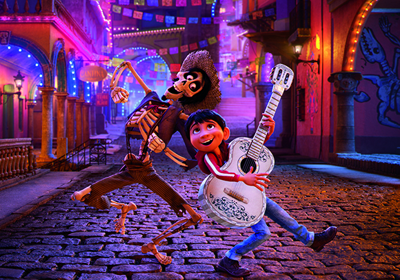 A skeleton and a young boy run joyfully down a cobblestone street decorated with colorful lights.