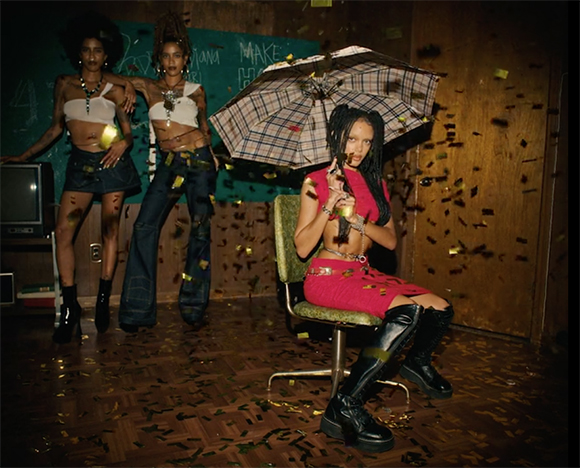 Three women in stylish outfits pose in a room as gold confetti falls around them.
