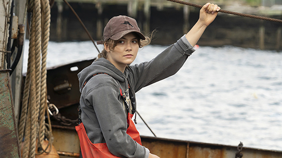 A young woman wearing a brown cap and grey hoodie holds a rope on a boat near the water.