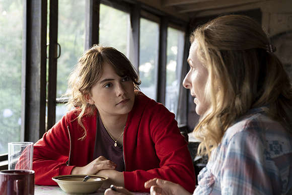 A young woman in a red hoodie looks up at a blonde woman talking to her in a room with large windows.