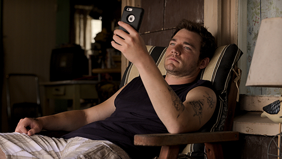 A man in a dark tank top reclines in a chair while holding a smartphone up to his face.