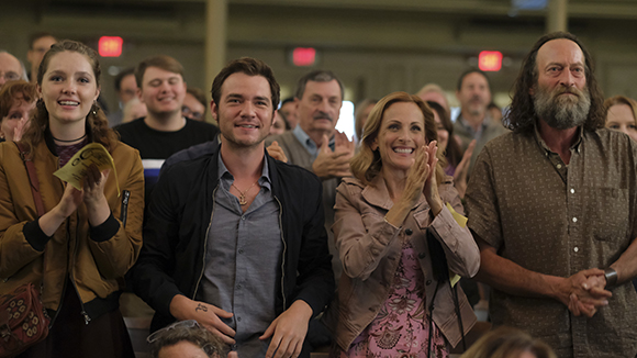 A diverse group of people smiling and clapping while sitting in an indoor audience.