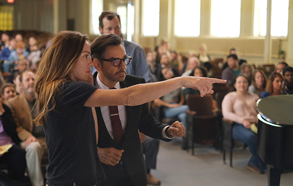 A woman points forward while a man in a suit and glasses stands next to her in a room filled with people.