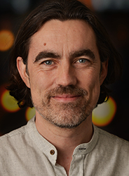 A man with dark hair, blue eyes, and a salt-and-pepper beard smiling against a blurred background.