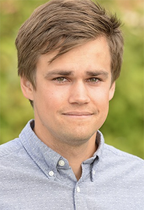A young man with brown hair and a light blue button-down shirt smiling slightly against a green background.