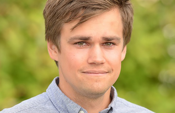 A young man with short brown hair and green eyes smiling slightly against a blurred green background.