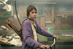 A young boy in a purple hoodie sits on a bicycle with a bow and quiver of arrows on his back.