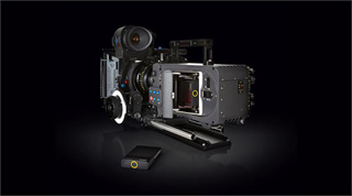 A complex professional cinema camera rig with various attachments and a small black accessory box on a dark surface.