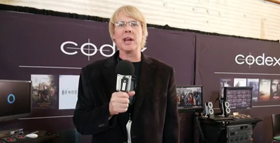 A man with blonde hair and glasses holds a microphone in front of a dark purple Codex branded backdrop.
