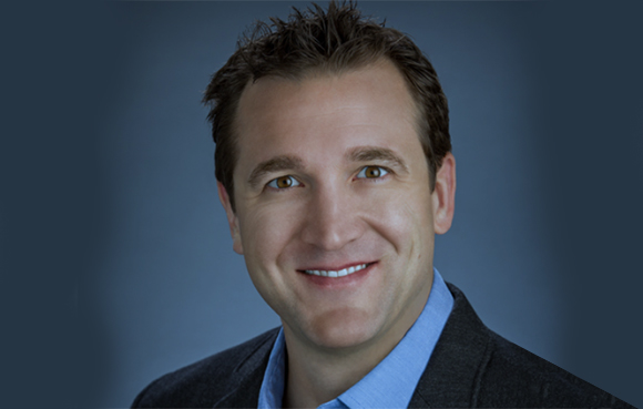A smiling man with short brown hair wearing a blue shirt and dark blazer against a blue background.