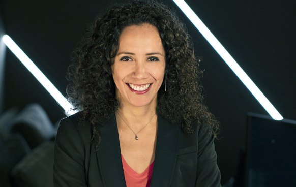 A smiling woman with dark curly hair wearing a black blazer and a red top.
