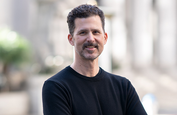 A man with dark curly hair and a mustache smiling in front of a blurred outdoor background.