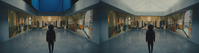 A person in a black bodysuit stands in a large room facing panoramic video projections of an ornate interior.