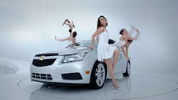 A woman in a white dress leans against a silver Chevrolet sedan with dancers blurred in the background.