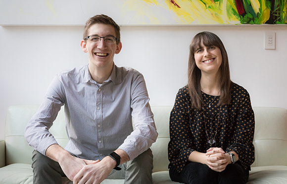A man in a grey shirt and a woman in a black patterned blouse sit smiling on a light sofa.